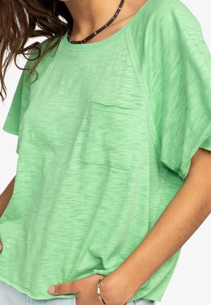 Femme portant un T-shirt vert clair à manches courtes avec une poche poitrine, un collier de perles et un bracelet argenté, main placée dans la poche d'un pantalon en jean clair.