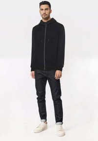 Uomo in piedi che indossa una felpa nera con zip, jeans scuri con risvolti e sneaker bianche su uno sfondo chiaro uniforme.