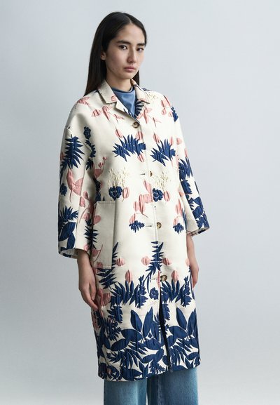 Manteau à motif floral avec tissu beige, comprenant des dessins botaniques bleus et roses. Conçu avec un col, des boutons et des poches.