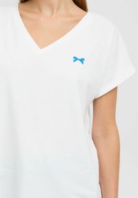 Tricou alb cu decolteu în V, fabricat din bumbac moale, având un mic broderie cu fundiță albastră pe partea stângă a pieptului și mâneci scurte răsucite.