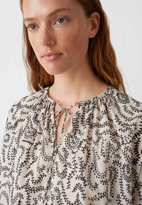 Blouse featuring a lightweight, cream fabric with black leaf-patterned embroidery. Gathered neckline with a tie detail.