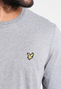 Grå bomullströja med rund halsringning och en svart och gul broderad fågel-logotyp på vänster bröst.