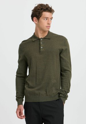 Man met krullend haar die een olijfgroen poloshirt met lange mouwen en een zwarte broek draagt, kijkt naar rechts tegen een effen achtergrond.