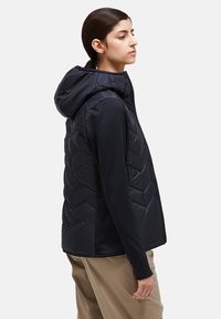 Black hooded puffer vest with a chevron quilted design. Soft, shiny fabric; full zip closure; paired with a long-sleeve top and beige pants.