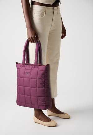Padded purple tote bag with quilted texture, dual handles, zipper closure, and a logo patch, held alongside light-colored pants.