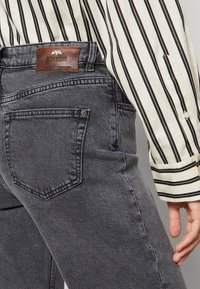 Jeans en denim gris avec une patch en cuir sur la taille, présentant un design à jambe droite, des coutures discrètes et un détail de poche arrière.