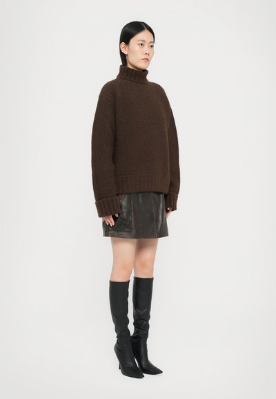 Brauner gestrickter Rollkragenpullover, oversize Schnitt, kombiniert mit einem schwarzen Lederrock und knielangen schwarzen Stiefeln mit Absatz. Glatte Textur.