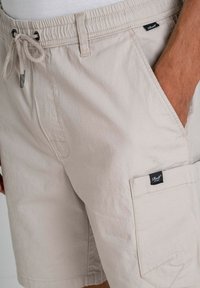 Pantalones cortos cargo beige hechos de tela ligera. Con cintura elástica ajustable, bolsillos laterales y una etiqueta en la pierna. Superficie texturizada.
