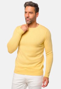 Pull en laine jaune tricoté, à manches longues et avec un col rond. Le mannequin porte un pantalon blanc, mettant en avant une silhouette ajustée.