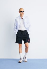 Kortærmet lyseblå skjorte med knapper, hvid tanktop, sorte shorts med rødt logo, hvide sokker og grå sneakers. Står op af en hvid væg.