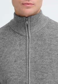 Grijze ritstrui met ribbeltextuur en hoge kraag. Voorzien van een voorrijt en zacht materiaal. Close-up van de bovenste helft.