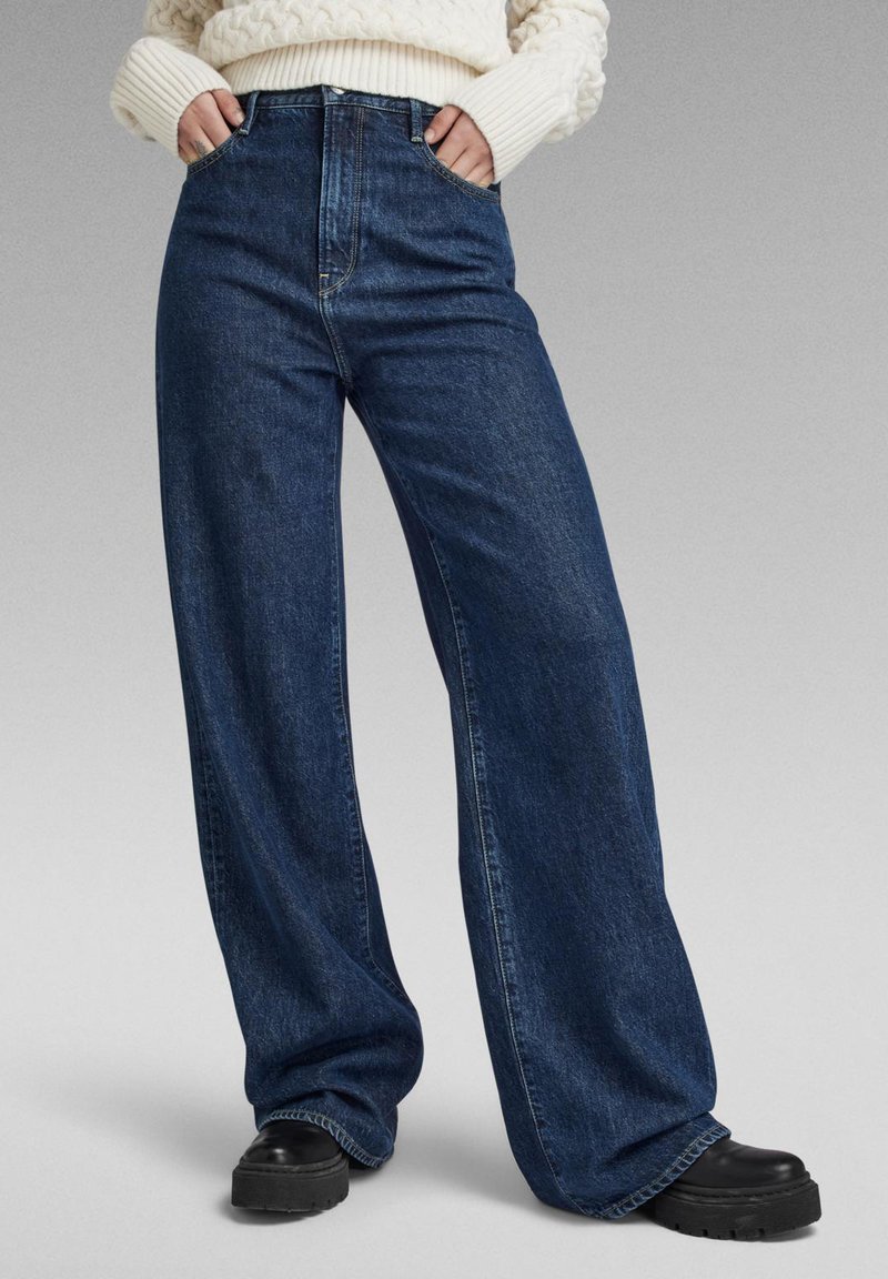 Jeans din denim întunecat, cu croială largă și talie înaltă, dotate cu buzunare frontale și o textură netedă, completate de botine negre cu talpă groasă.