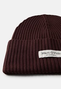 Gorro de punto burdeos con textura acanalada. Presenta una etiqueta blanca con texto negro, el nombre de la marca "Marc O'Polo" y "EST. EN STOCKHOLM."