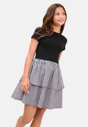 Fată purtând un top negru cu mâneci scurte și o fustă stratificată în carouri alb-negru, privind în lateral cu un zâmbet ușor.