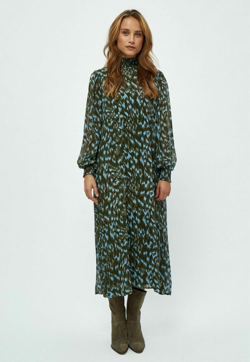 Langes, fließendes Kleid in grün-blauem abstraktem Muster, hoher Kragen, lange Ärmel mit elastischen Bündchen, kombiniert mit olivgrünen, knielangen Stiefeln.