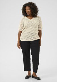 Vrouw met krullend haar, gekleed in een beige gebreide korte mouw trui, zwarte broek tot de enkels en zwarte ballerina's, staand voor een witte achtergrond.