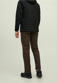 Chaqueta negra con capucha, parte frontal acolchada y mangas lisas, combinada con pantalones marrones y zapatillas negras, vista desde atrás sobre un fondo claro.