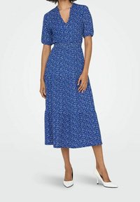 Sininen midi-mekko valkoisilla kukkakuvioilla, lyhyet puffihihat, V-mallinen kaula-aukko ja kerroksellinen helma. Yhdistetty valkoisiin, teräväkärkisiin korkokenkiin.