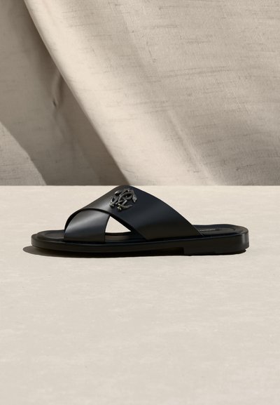 Μαύρο δερμάτινο σανδάλι με λουράκια που διασταυρώνονται και ασημί διακοσμητικό έμβλημα, απεικονίζεται πάνω σε ομαλή μπεζ επιφάνεια με φόντο από κρεμασμένο ύφασμα.