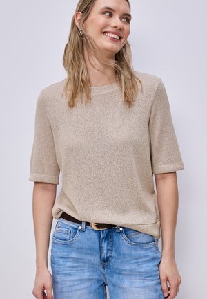 Femme souriante aux cheveux blonds mi-longs portant un haut beige en maille à manches courtes, rentré dans un jean bleu clair avec une ceinture noire.