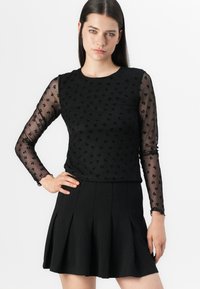 Čierny top s dlhým rukávom z texturovanej čiernej tkaniny so kvetinovými vzormi, v kombinácii s plisovanou čiernou sukňou. Obe sú elegantné a prispôsobené postave.