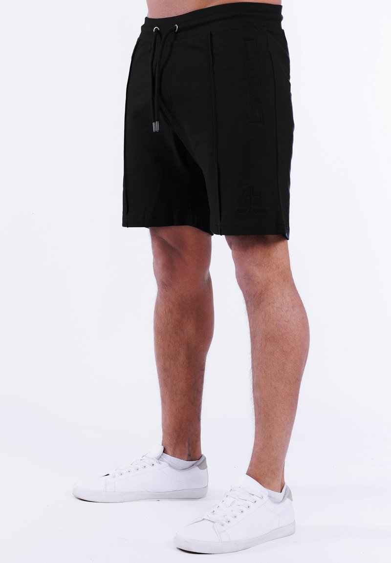 Schwarze Shorts mit einem Kordelzugbund, Seitentaschen und einem geprägten Logo am unteren Bein. Hergestellt aus einem glatten, leichten Stoff.