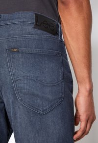 Homme portant un jean Lee bleu foncé, montrant la poche arrière et la boucle en cuir de la marque, avec le bras droit détendu le long du corps.