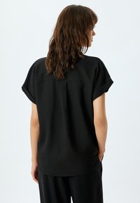 Blusa negra de manga corta con ajuste relajado, tejido texturizado y detalle de yugo en la parte posterior. El dobladillo redondeado y los puños ofrecen un diseño sencillo.