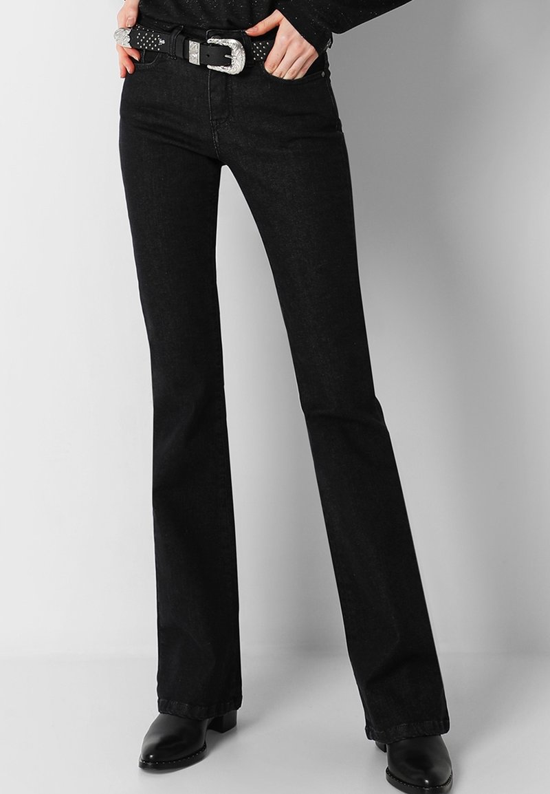 Jean flare en denim noir avec une texture lisse, taille mi-haute et boucle de ceinture à clous argentés. Comprend un design classique à cinq poches.