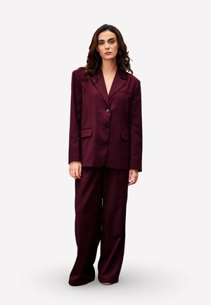 Vrouw met lange krullende haren, gekleed in een losse donker burgundy suit met wijde broekspijpen, staand tegen een effen lichte achtergrond.