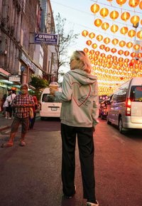 Felpa grigia chiara con design a cuore rosso, pantaloni neri, che tiene una bevanda, in piedi su una strada con lanterne arancioni sopra.