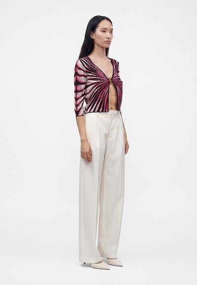 Femme aux longs cheveux noirs porte un cardigan court rose et noir à motifs, un pantalon blanc large taille haute et des chaussures blanches pointues.