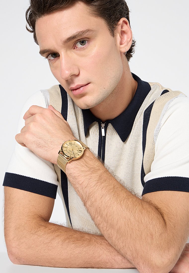 Polo en maille à manches courtes et col, crème et marine, avec une fermeture éclair à l'avant. Une montre dorée avec un bracelet en mailles repose sur une main.