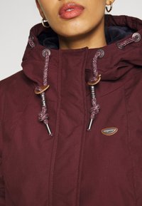 Femme portant une veste à capuche bordeaux avec des cordons tressés et un petit patch logo sur la poitrine, lèvres visibles, portant des boucles d'oreilles en or.