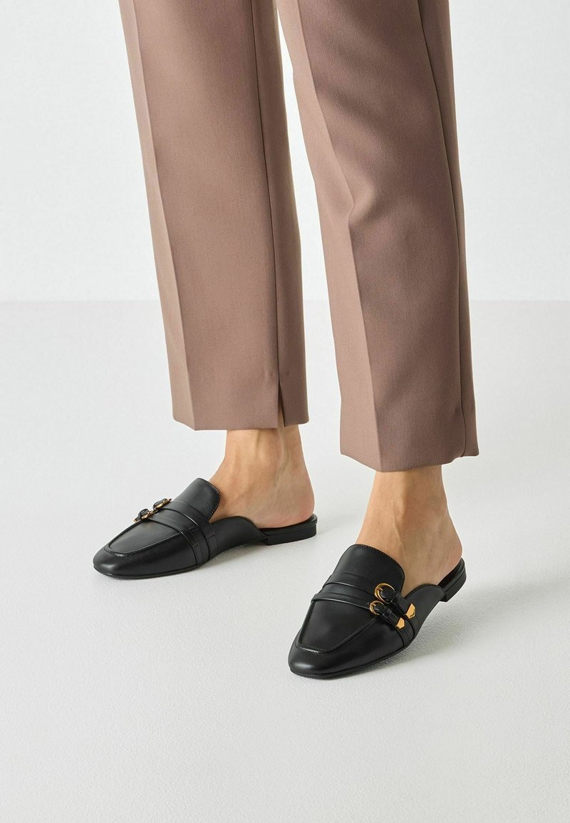 Personne portant un pantalon marron court et des mocassins en cuir noir sans talon avec des détails de boucle dorée, debout sur un sol clair.
