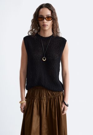 Femme aux cheveux ondulés, portant des lunettes de soleil marron, un haut en maille noir sans manches, une jupe marron, des bijoux en or et un collier pendentif, de face.
