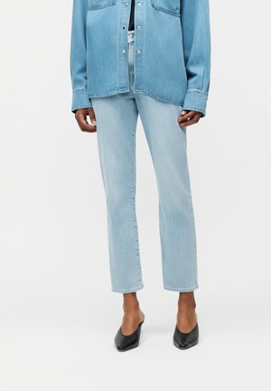 Personne portant un jean droit bleu clair, une chemise en denim assortie et des mules à talons noirs à bout pointu sur fond blanc.