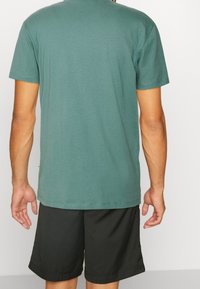 T-shirt à manches courtes et col rond en tissu vert doux, associé à un short noir. Les détails des coutures et la finition de l'ourlet sont visibles.