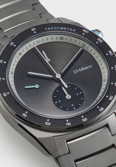 Rund grå metalur med en tachymeterskala, sort urskive, sølvfarvede timemarkører og en børstet finish på kassen og armbåndet.