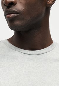 Κοντινό πλάνο του κάτω μέρους προσώπου και του λαιμού ενός άνδρα με σκουρόχρωμο δέρμα, που φοράει ένα ανοιχτό γκρι μπλουζάκι με στρογγυλή λαιμόκοψη σε απλό φόντο.