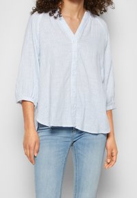 Kobieta ubrana w luźną bluzkę w pionowe, jasnoniebiesko-białe paski z guzikami oraz jasnoniebieskie dżinsy, stojąca na prostym tle.