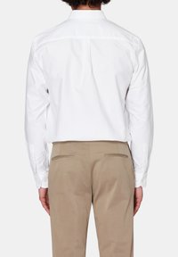 Camicia bianca a maniche lunghe con colletto piccolo, piega sul retro e polsini con bottone, abbinata a pantaloni beige con due tasche posteriori.