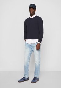 Marinefarbener Sweatshirt, hellrosa Hemd mit Kragen, hellblaue ausgewaschene Jeans, blaue Wildledersneaker und eine dunkelblaue Mütze mit "BOSS"-Logo.