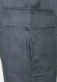 Mörkblått, strukturerat tyg med ett diagonalt mönster; har en framficka med lock och sömnadsdetaljer. Närbild av materialet.