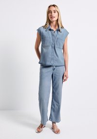 Donna in piedi contro uno sfondo bianco, che indossa una camicia in denim senza maniche di un leggero blu, pantaloni larghi abbinati e sandali bianchi con strappi.