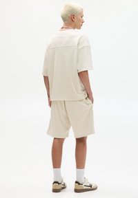 Lichtbeige, kortgemouwen netshirt in combinatie met bijpassende shorts, met een relaxte pasvorm en zijzakken. Draagt witte sokken en sneakers.