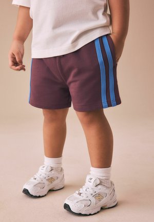 Väikelaps kannab valgeid tenniseid, valgeid sokke, bordoopunaseid lühikesi pükse siniste triipudega ja valget särki, seisab beežil põrandal.