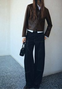 Brązowa skórzana krótka kurtka na białej koszuli, w połączeniu z szerokimi ciemnoniebieskimi dżinsami z widocznym przeszyciem, trzymając małą czarną torebkę.