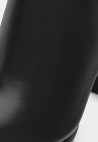 Μαύρο δερμάτινο μποτάκι μέχρι τον αστράγαλο με γυαλιστερή φινίρισμα, κωνικό σχήμα, λεία υφή και μινιμαλιστικό σχέδιο, χωρίς ορατές διακοσμήσεις.
