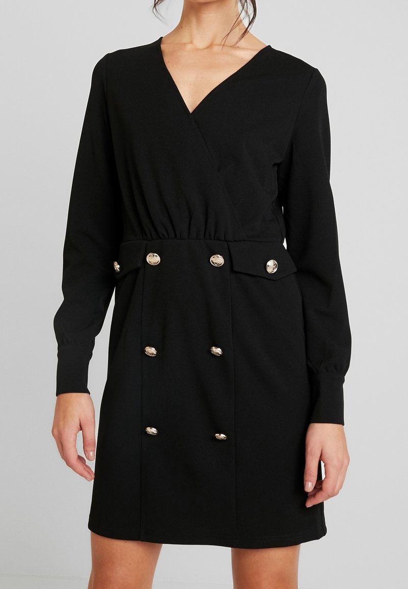 Femme portant une robe portefeuille noire à manches longues avec des boutons dorés disposés en deux rangées verticales sur la partie jupe.